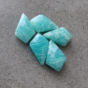 May include: Five teal green gemstone chips with a faceted, irregular shape. The chips are arranged in a cluster on a gray textured surface.