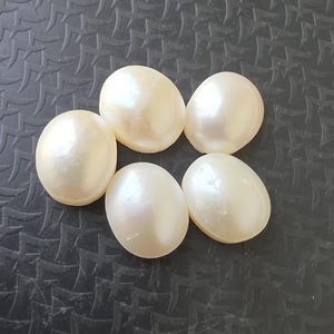 Op de afbeelding: Zes cr&egrave;mekleurige, ovale parels liggen op een gestructureerd, donkergrijs oppervlak. De parels hebben een gladde, iriserende glans die het licht reflecteert. Ze kunnen worden gebruikt voor het maken van sieraden of andere knutselprojecten.