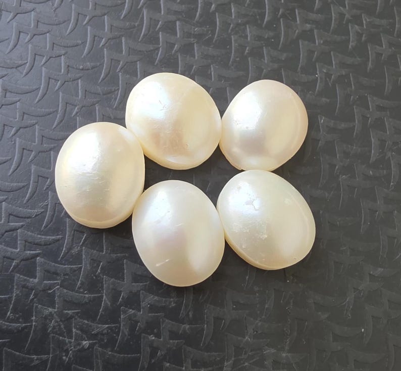 Op de afbeelding: Zes cr&egrave;mekleurige, ovale parels liggen op een gestructureerd, donkergrijs oppervlak. De parels hebben een gladde, iriserende glans die het licht reflecteert. Ze kunnen worden gebruikt voor het maken van sieraden of andere knutselprojecten.