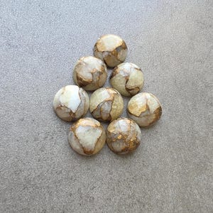 May include: Eight round, polished stones with a cream and gold color scheme. The stones have a marbled appearance with gold veining throughout. The stones are arranged in a triangular shape on a gray surface.