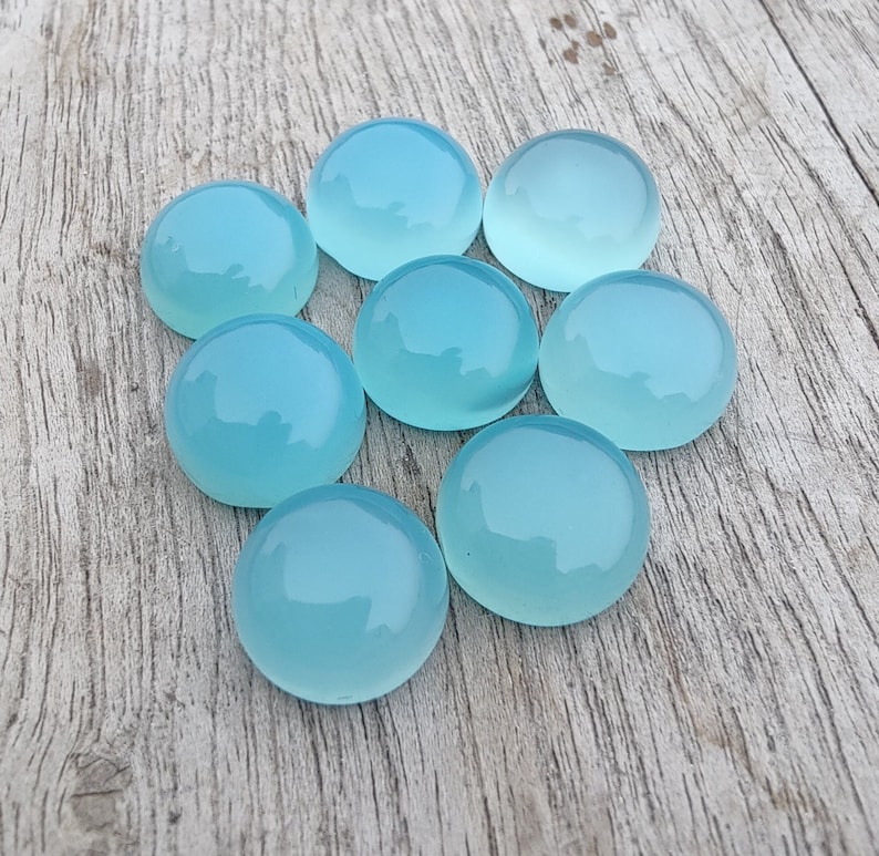 Pu&ograve; includere: Otto pietre preziose rotonde, lisce e di colore azzurro chiaro. Le pietre preziose sono disposte in cerchio su una superficie di legno marrone chiaro.