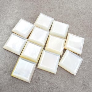 May include: A collection of ten square-shaped buttons with a smooth, iridescent white finish. Each button features a bevelled edge and a flat centre, displaying a pearlescent sheen that reflects light across the surface of the square components.