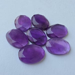 May include: Seven faceted, oval-shaped amethyst gemstones in varying shades of purple are arranged on a light surface. The gemstones have a polished, reflective surface, and the facets catch the light, creating a sparkling effect. These gemstones are suitable for jewelry making.