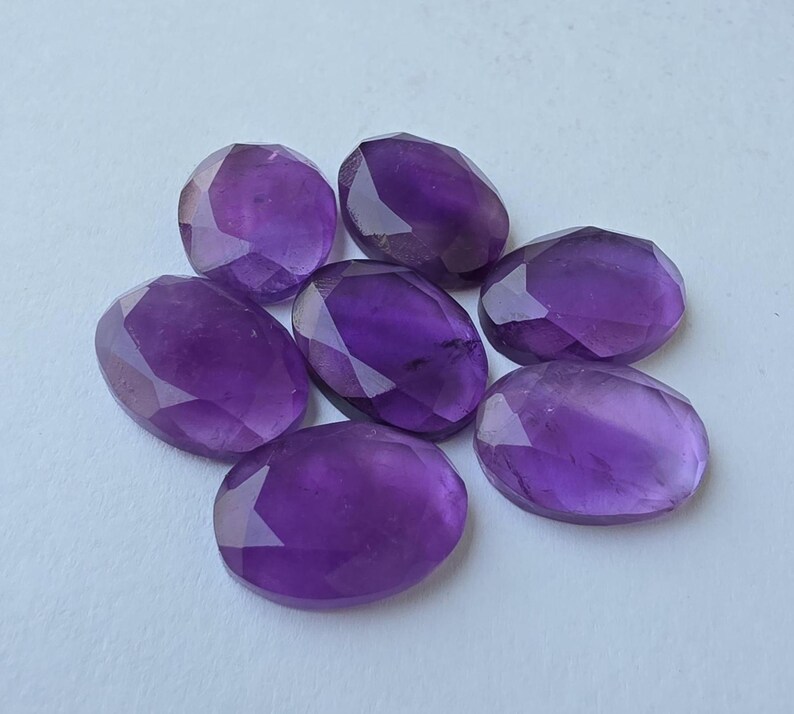 May include: Seven faceted, oval-shaped amethyst gemstones in varying shades of purple are arranged on a light surface. The gemstones have a polished, reflective surface, and the facets catch the light, creating a sparkling effect. These gemstones are suitable for jewelry making.