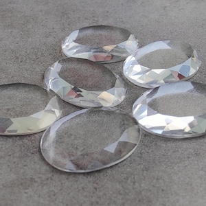 May include: Six clear, faceted oval-shaped gemstones are arranged on a gray surface. The gems have a sparkling, reflective quality, with multiple facets catching the light. The overall effect is one of elegance and sparkle.