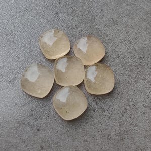 May include: Six light amber-colored, square-shaped gemstones with rounded corners are arranged in a circular pattern. The stones have a translucent quality, reflecting light. The background is a textured gray surface.