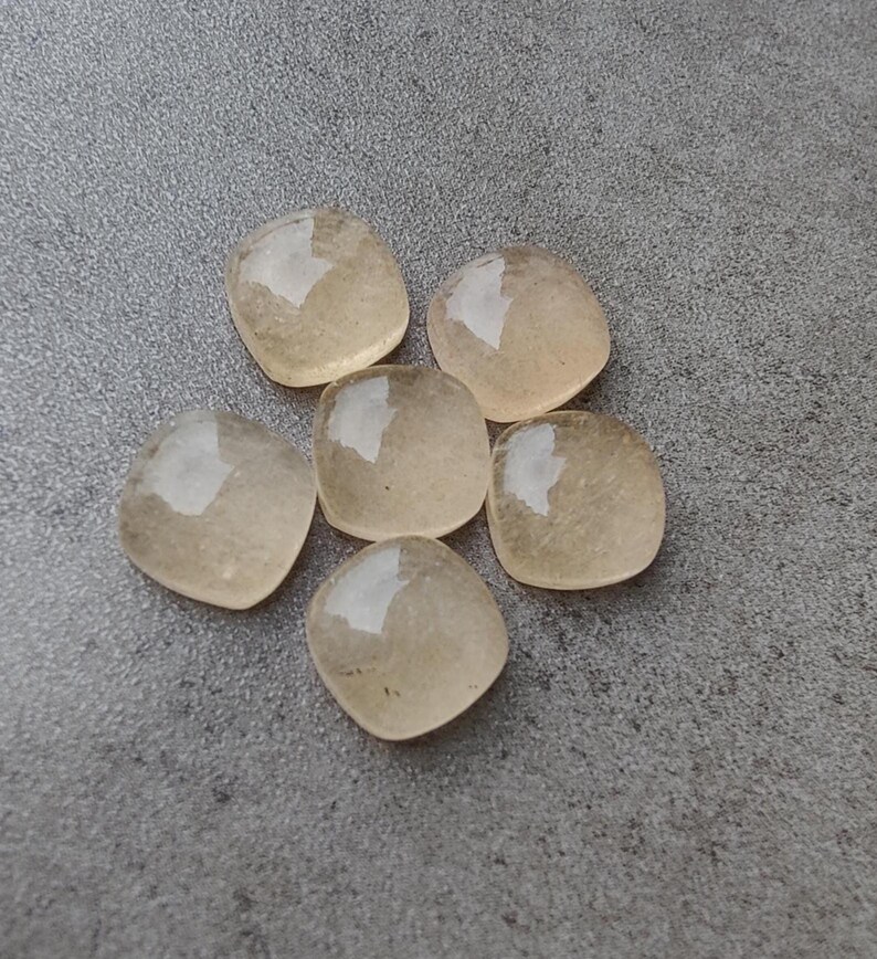 May include: Six light amber-colored, square-shaped gemstones with rounded corners are arranged in a circular pattern. The stones have a translucent quality, reflecting light. The background is a textured gray surface.