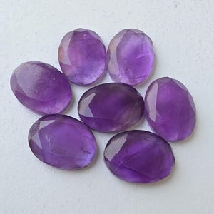 May include: Seven faceted, oval-shaped amethyst gemstones. The gemstones are a deep purple color and have a polished, reflective surface. The facets catch the light, creating a sparkling effect. The gemstones are arranged in a circular pattern.