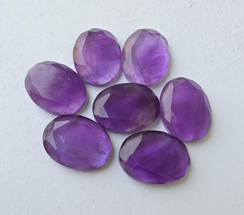 May include: Seven faceted, oval-shaped amethyst gemstones. The gemstones are a deep purple color and have a polished, reflective surface. The facets catch the light, creating a sparkling effect. The gemstones are arranged in a circular pattern.