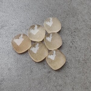 May include: Six translucent, square-shaped beads with rounded corners. Each bead has a light brown hue with a white, heart-shaped design in the center. The beads are arranged on a gray surface.