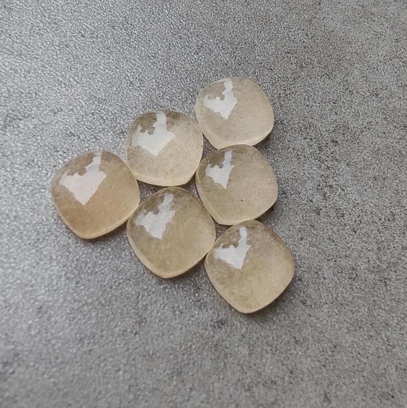 May include: Six translucent, square-shaped beads with rounded corners. Each bead has a light brown hue with a white, heart-shaped design in the center. The beads are arranged on a gray surface.