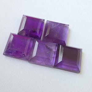 Peut inclure: Cinq pierres pr&eacute;cieuses d'am&eacute;thyste taill&eacute;es en carr&eacute;, de diff&eacute;rentes nuances de violet. Les pierres sont dispos&eacute;es sur une surface blanche, mettant en valeur leurs facettes et variations de couleur. Id&eacute;al pour la joaillerie.