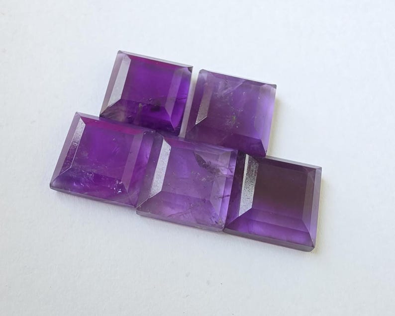 Peut inclure: Cinq pierres pr&eacute;cieuses d'am&eacute;thyste taill&eacute;es en carr&eacute;, de diff&eacute;rentes nuances de violet. Les pierres sont dispos&eacute;es sur une surface blanche, mettant en valeur leurs facettes et variations de couleur. Id&eacute;al pour la joaillerie.