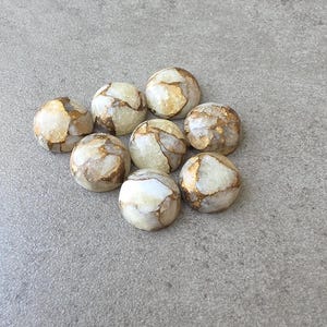 May include: Nine round, polished stones with a cream and gold marbled pattern. The stones are arranged in a loose cluster on a gray surface. The gold veining adds a touch of elegance to the natural stone.