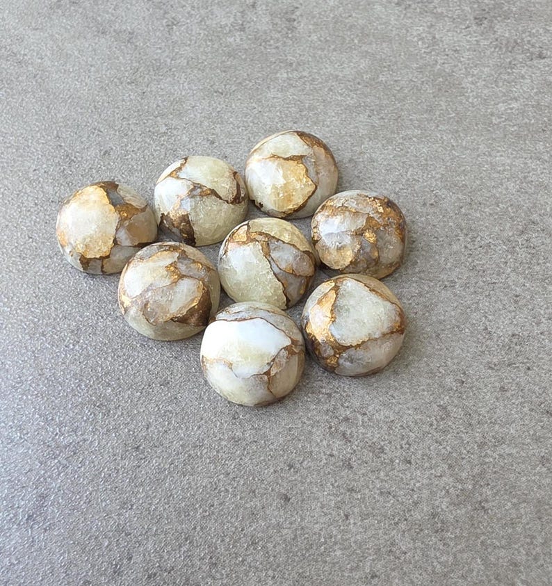 May include: Nine round, polished stones with a cream and gold marbled pattern. The stones are arranged in a loose cluster on a gray surface. The gold veining adds a touch of elegance to the natural stone.