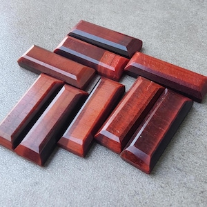 May include: A collection of eight red tiger's eye gemstone cabochons, each with a rectangular shape and polished surface. The stones are arranged in a staggered pattern, showcasing their rich red color and subtle chatoyancy.