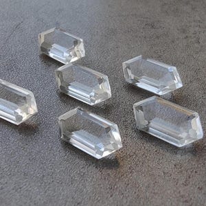 May include: Six clear, faceted, octagonal-shaped gemstones. The gemstones are arranged in a semi-circle on a gray surface.