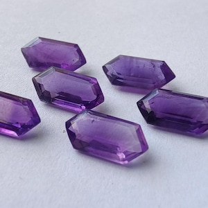 May include: Six faceted, hexagonal amethyst gemstones in varying shades of purple are scattered on a white surface. The gemstones have a polished, crystalline appearance, reflecting light.