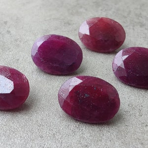 Puede incluir: Cinco piedras preciosas de rubí de forma ovalada y facetadas en varios tonos de rojo. Las piedras preciosas están dispuestas en semicírculo sobre una superficie gris.