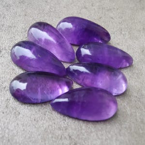 Op de afbeelding: Een groep van zeven paarse amethist edelsteen cabochons, elk gevormd als een traan, met een glad, gepolijst oppervlak.