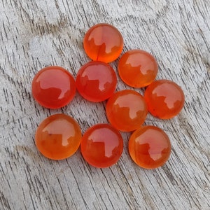 May include: Nine round, orange carnelian cabochons, polished and smooth, arranged in a triangular pattern on a wooden surface.