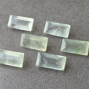 Puede incluir: Seis gemas rectangulares de color verde pálido con bordes facetados están dispuestas sobre una superficie gris. Las gemas tienen una cualidad translúcida, con sutiles variaciones de color y claridad. La estética general es limpia y minimalista.