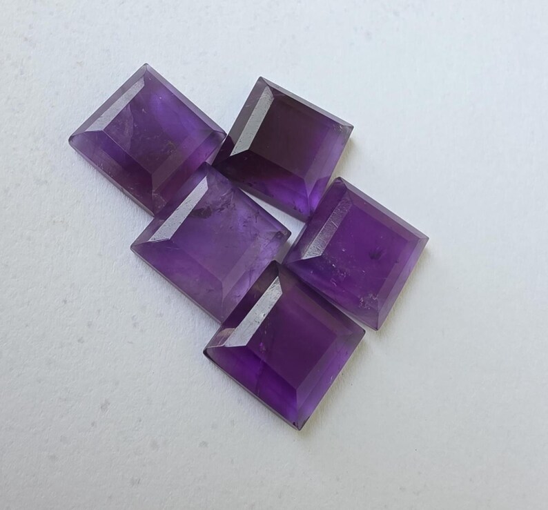 Peut inclure: Cinq pierres pr&eacute;cieuses d'am&eacute;thyste taill&eacute;es en carr&eacute;. Chaque gemme pr&eacute;sente une teinte violette profonde et une surface facett&eacute;e, captant la lumi&egrave;re. Les pierres sont dispos&eacute;es sur une surface claire, mettant en valeur leur couleur et leur taille.