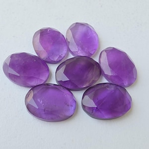 May include: Seven faceted oval amethyst gemstones in varying shades of purple are arranged on a white surface. The gemstones have a polished, reflective surface, showcasing their cut and clarity. The arrangement is circular.