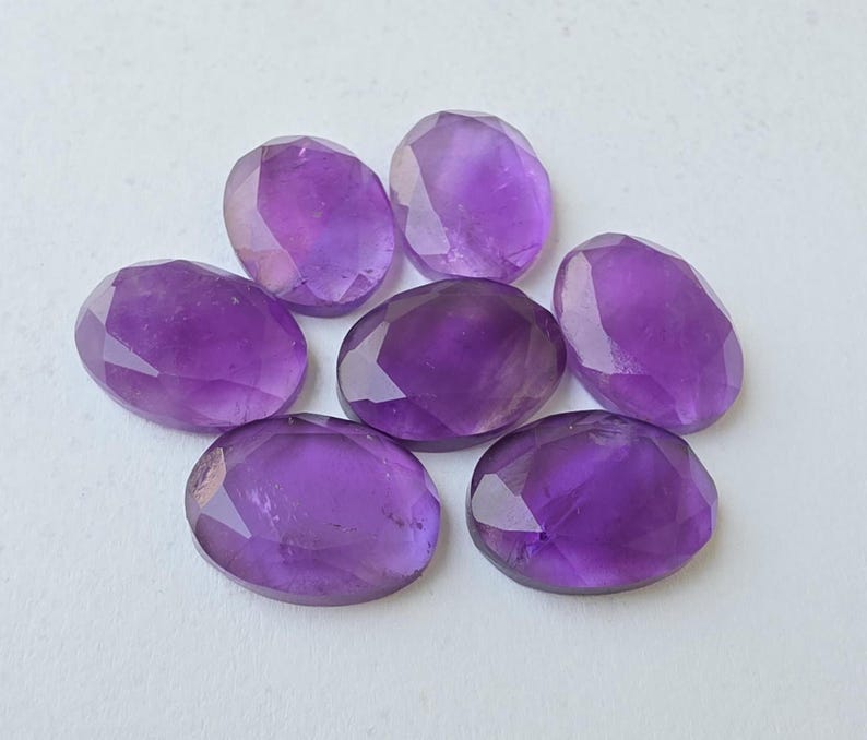 May include: Seven faceted oval amethyst gemstones in varying shades of purple are arranged on a white surface. The gemstones have a polished, reflective surface, showcasing their cut and clarity. The arrangement is circular.