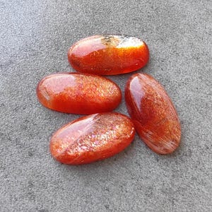 K&ouml;nnte beinhalten: Vier ovale Sonnenstein-Cabochons in Orange- und Rott&ouml;nen mit irisierendem Schimmer. Die Steine sind glatt und poliert.
