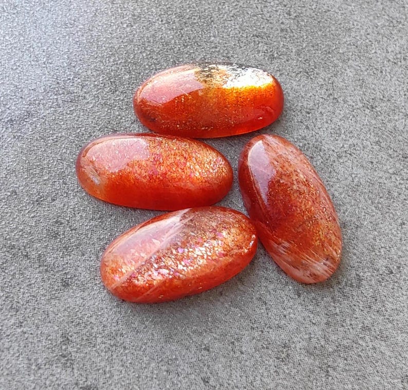 K&ouml;nnte beinhalten: Vier ovale Sonnenstein-Cabochons in Orange- und Rott&ouml;nen mit irisierendem Schimmer. Die Steine sind glatt und poliert.