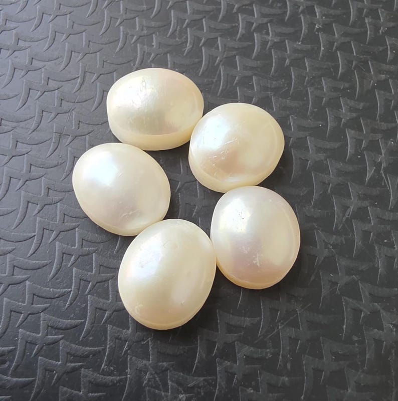 Op de afbeelding: Vijf ovale, cr&egrave;mekleurige parels met een iriserende glans, geplaatst op een donkergrijze, getextureerde achtergrond. De parels hebben een glad oppervlak en reflecteren het licht. Geschikt voor het maken van sieraden of andere decoratieve items.