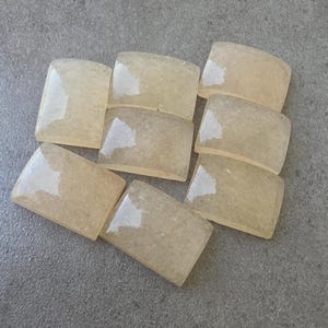 May include: A collection of nine rectangular, light yellow-beige gemstones with rounded corners. The stones have a translucent quality and a slightly textured appearance, arranged on a gray surface. The gemstones are likely for jewelry making or crafting.