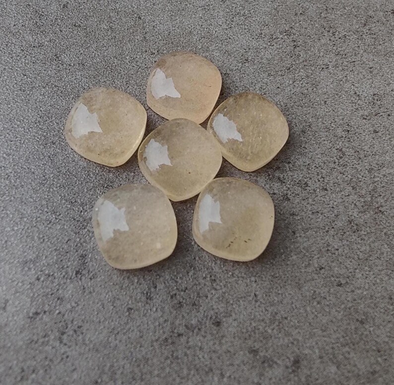 May include: Six light yellow, square-shaped gemstones with rounded corners are arranged on a gray surface. Each stone has a glossy, reflective surface, catching the light. The gemstones appear translucent.