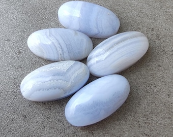May include: Five oval-shaped blue lace agate stones with white and light blue banding. The polished gemstones are arranged on a gray surface, showcasing their natural patterns and colors. These stones are often used in jewelry and crystal healing.