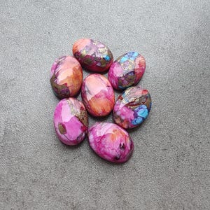May include: Seven oval-shaped gemstones in shades of pink, orange, and blue with gold accents. The gemstones are arranged in a circular pattern against a gray background. The stones have a polished, smooth surface.