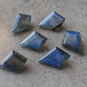 May include: Six faceted labradorite gemstones in a trapezoid shape are shown. The stones display blue, grey, and green colours, with internal patterns. The gemstones are arranged on a grey surface.