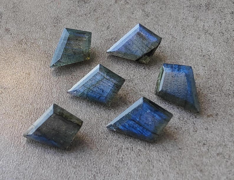 May include: Six faceted labradorite gemstones in a trapezoid shape are shown. The stones display blue, grey, and green colours, with internal patterns. The gemstones are arranged on a grey surface.