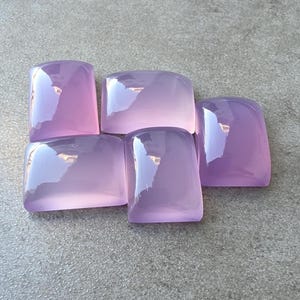 Pu&ograve; includere: Cinque pietre preziose rettangolari lavanda lucide con angoli arrotondati sono disposte su una superficie grigia. Le pietre hanno una qualit&agrave; traslucida, riflettendo la luce e creando riflessi. Le pietre preziose sono probabilmente per la creazione di gioielli o la collezione.