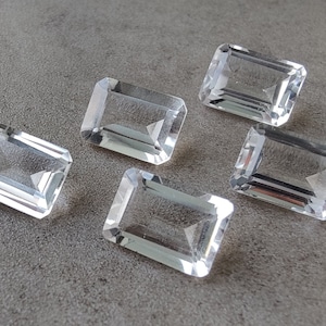 Pode incluir: Seis gemas transparentes, retangulares e facetadas. As gemas estão dispostas em semicírculo sobre uma superfície cinza.