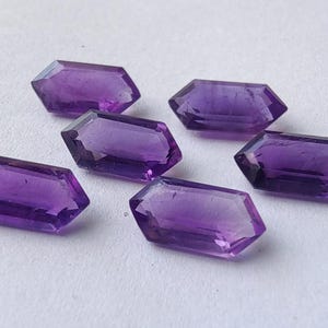 K&ouml;nnte beinhalten: Sechs facettierte Amethyst-Edelsteine ​​mit tiefvioletter Farbe sind auf einer wei&szlig;en Oberfl&auml;che angeordnet. Jeder Edelstein hat eine rechteckige Form mit abgewinkelten Seiten und einer polierten, reflektierenden Oberfl&auml;che.