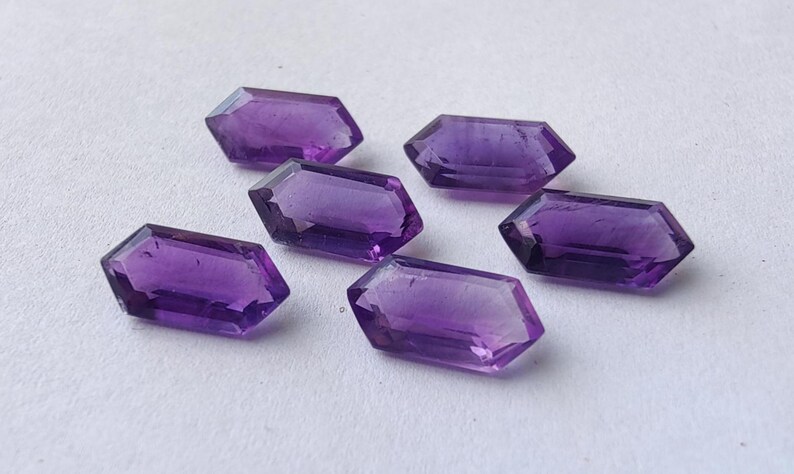 K&ouml;nnte beinhalten: Sechs facettierte Amethyst-Edelsteine ​​mit tiefvioletter Farbe sind auf einer wei&szlig;en Oberfl&auml;che angeordnet. Jeder Edelstein hat eine rechteckige Form mit abgewinkelten Seiten und einer polierten, reflektierenden Oberfl&auml;che.
