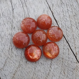 May include: Seven round, orange sunstone cabochons with a glittery, iridescent finish. The stones are arranged in a cluster on a light brown wooden surface.