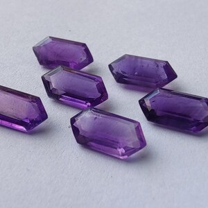 K&ouml;nnte beinhalten: Sechs facettierte, sechseckige Amethyst-Edelsteine ​​werden auf einer wei&szlig;en Oberfl&auml;che gezeigt. Die Edelsteine ​​sind tiefviolett und haben eine polierte, reflektierende Oberfl&auml;che. Die Edelsteine ​​sind mit mehreren Facetten geschliffen.
