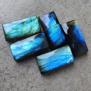 May include: Five rectangular labradorite stones with iridescent blue, green, and gold hues. The polished stones display a play of color, with flashes of light across their surfaces. The stones are arranged on a textured gray surface.