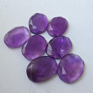 May include: Seven faceted oval amethyst gemstones in varying shades of purple. The gemstones are arranged on a white surface, showcasing their polished surfaces and internal clarity. Ideal for jewelry making or collecting.