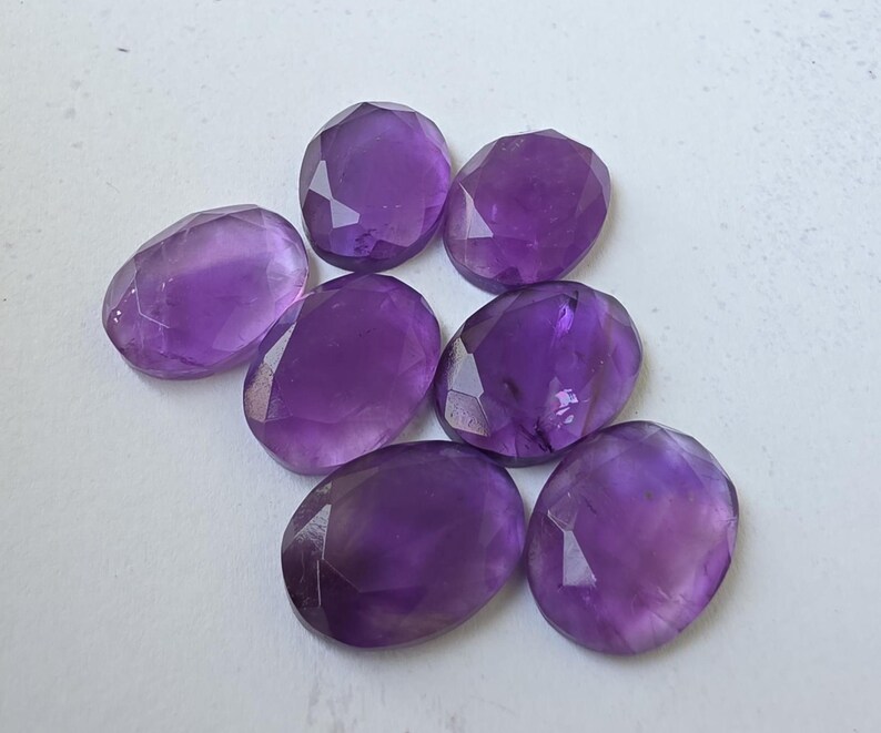 May include: Seven faceted oval amethyst gemstones in varying shades of purple. The gemstones are arranged on a white surface, showcasing their polished surfaces and internal clarity. Ideal for jewelry making or collecting.