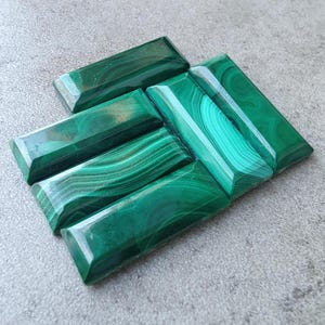 Puede incluir: Seis cabujones de malaquita verde, cortados en forma rectangular, con diferentes patrones de vetas verdes y blancas. Los cabujones están dispuestos en forma escalonada, con el cabujón más grande en la parte superior.
