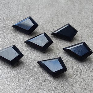 May include: Six dark blue, geometric-shaped gemstones with angled edges are arranged on a textured, gray surface. The gemstones have a polished, reflective surface, catching the light. The overall composition suggests a collection of loose gemstones.