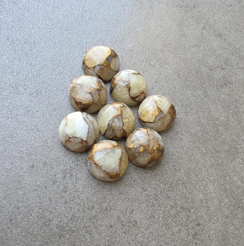 May include: Eight round, polished stones with a mix of white, tan, and gold colors. The stones have a marbled appearance with gold veining throughout. They are arranged in a loose cluster on a gray surface.